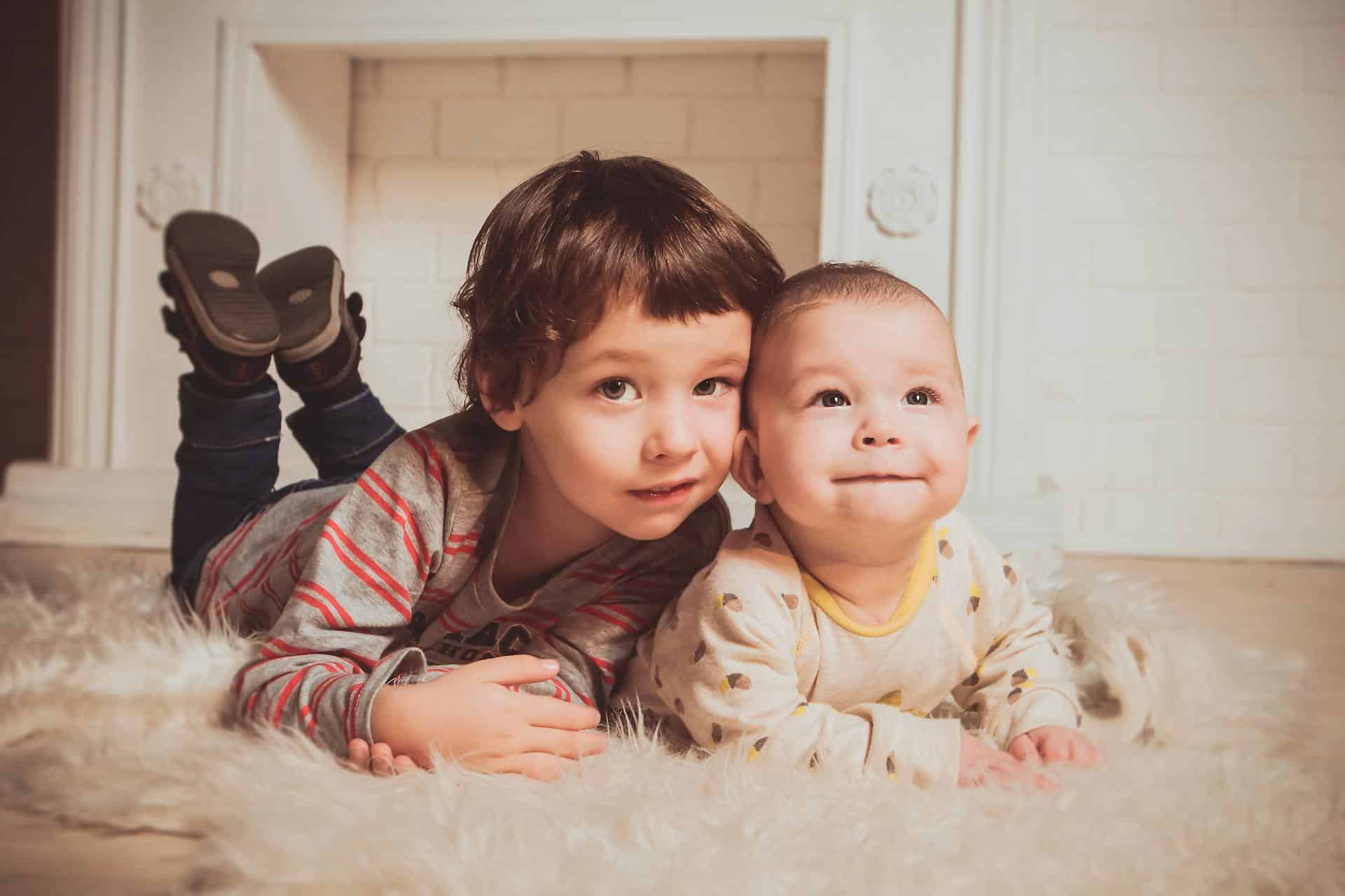 Older brother lying on a soft rug beside his baby sibling, both looking forward with warm expressions.