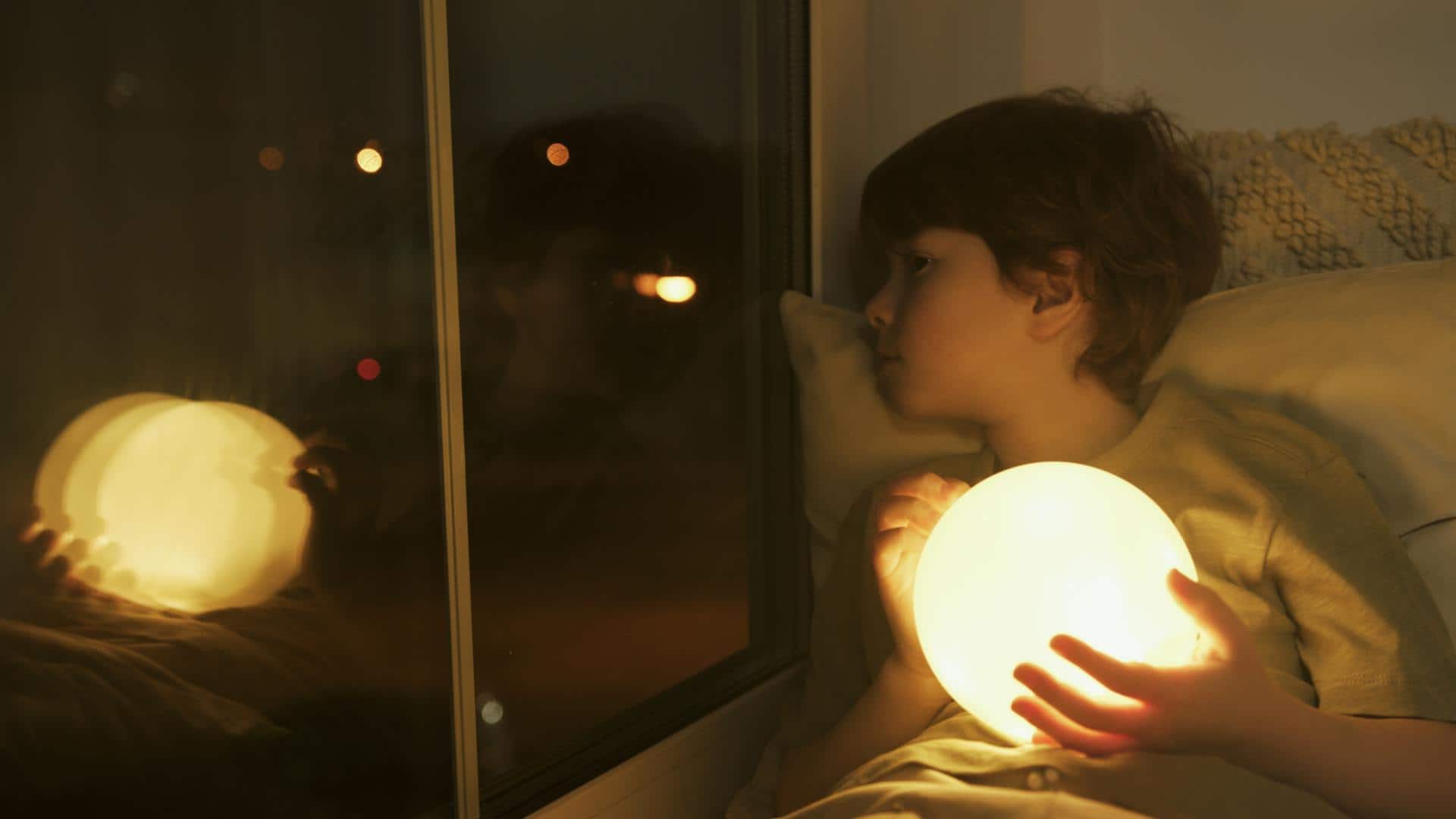 Young boy lying in bed at night holding a glowing light, staring out the window with a restless expression