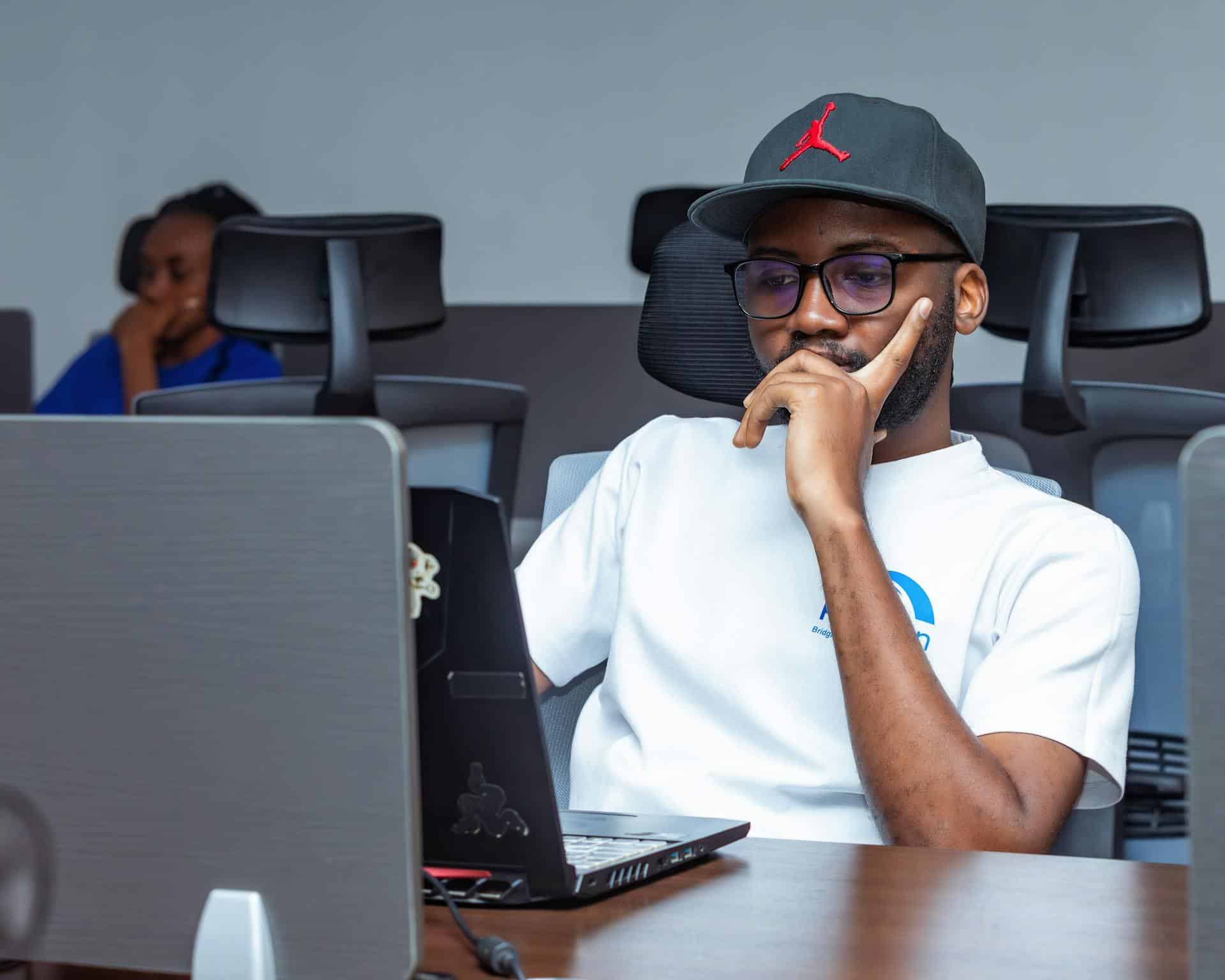 Adult male wearing a cap and glasses, seated in an office, looking deep in thought while using a laptop illustrating focus challenges common in ADHD in adult males.