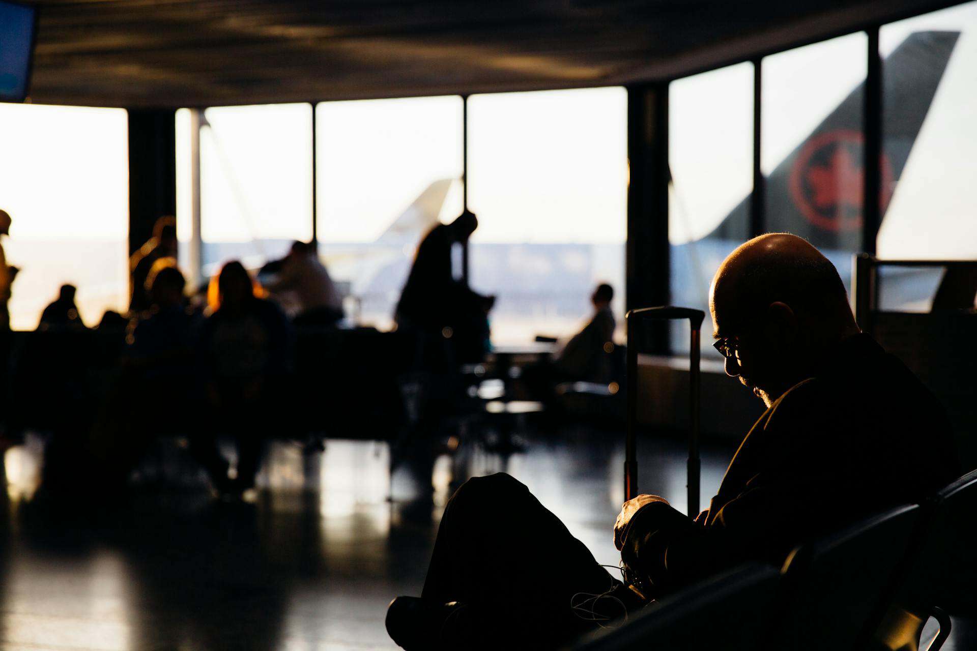 Travelers seated and waiting in an airport terminal, with warm sunlight casting shadows, reflecting the quiet tension and anticipation before a flight.