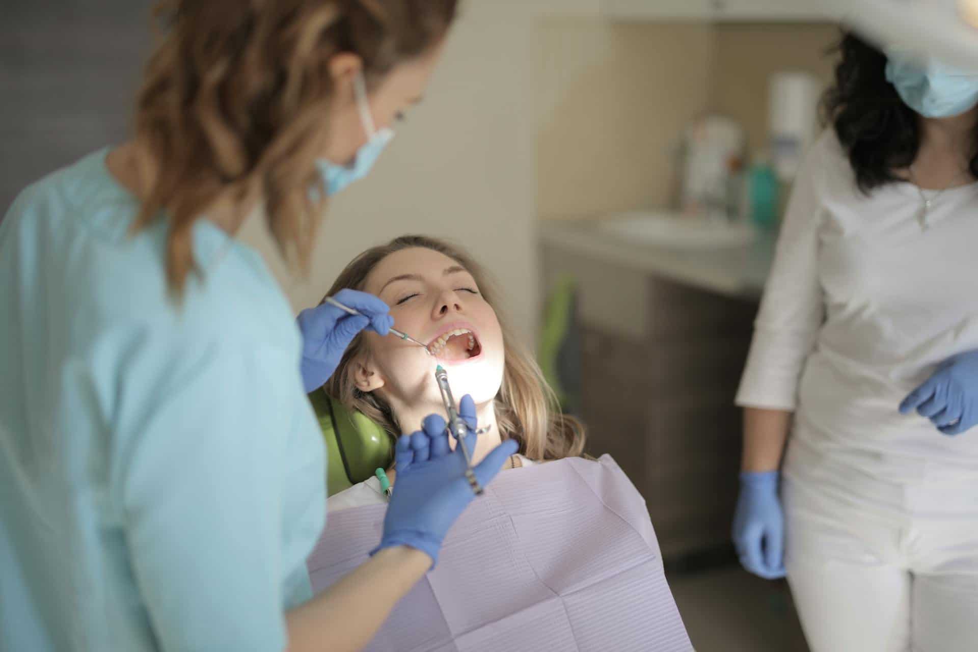 Female dentist examining a young woman’s open mouth during a dental checkup, with an assistant observing in the background.