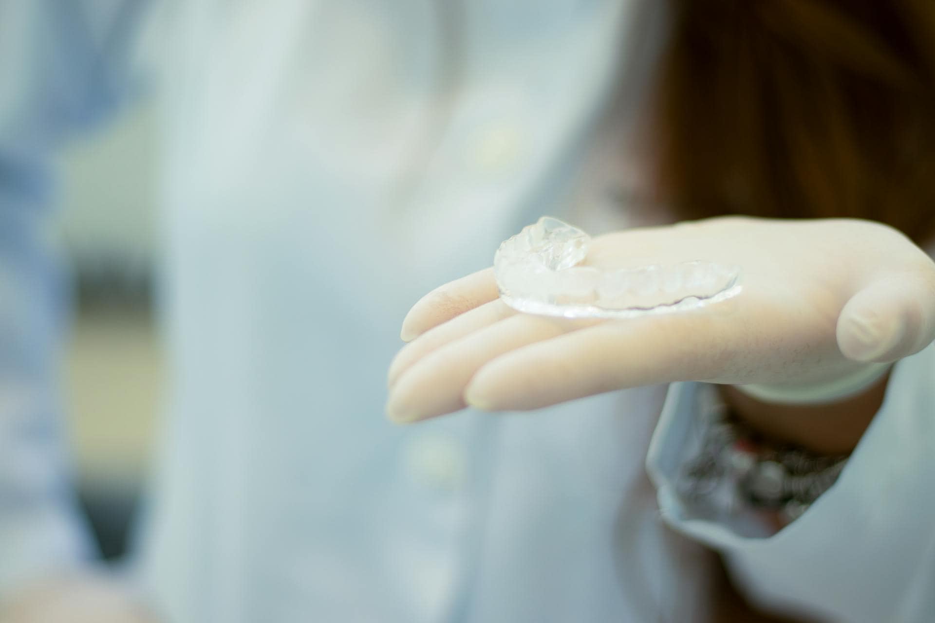 A close-up of a gloved hand holding a clear dental mouthguard, with a blurred background of a person in a lab coat.
