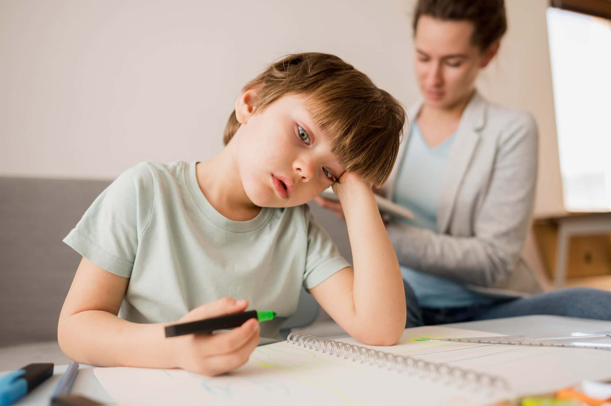 A young boy sitting at a table with a notebook, holding a marker and looking tired while an adult supports him in the background, symbolizing challenges of Dyspraxia in learning.
