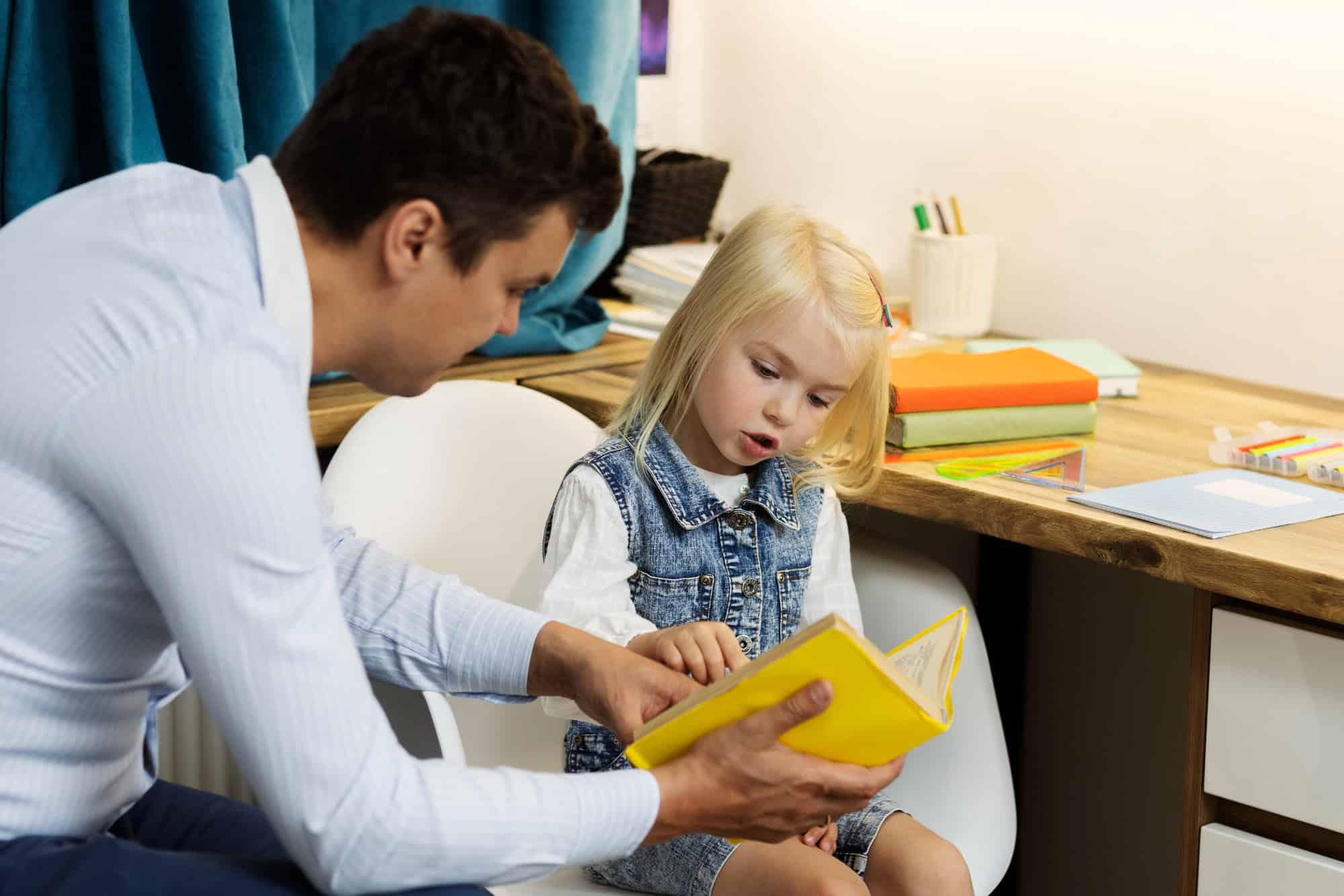 A young girl reads from a book with the guidance of an adult, symbolizing supportive learning strategies for children with Dyspraxia.