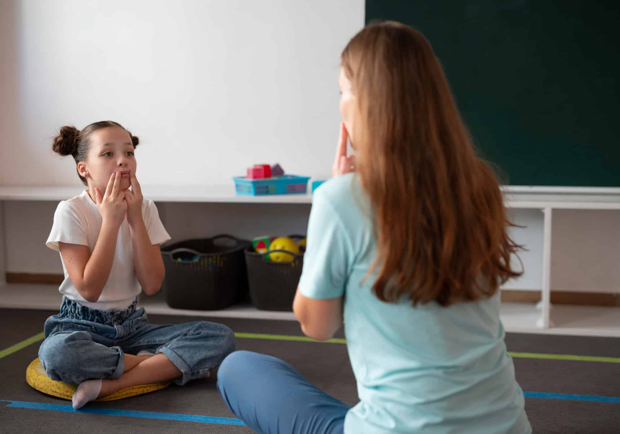 A young girl sits on the floor mimicking mouth movements during a speech therapy session with her instructor, illustrating supportive therapy for Dyspraxia.