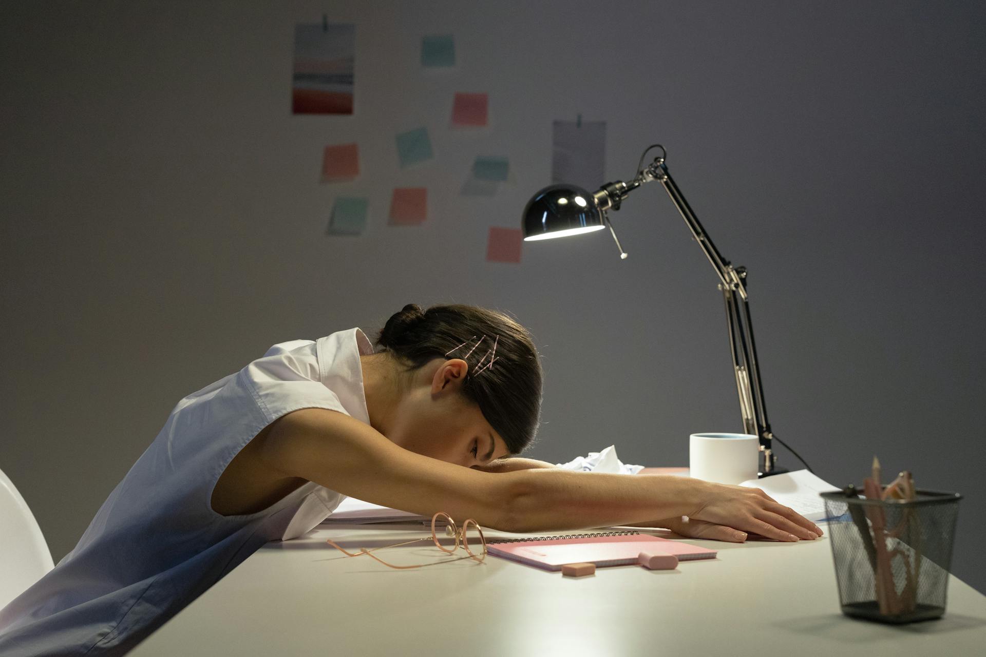 A young woman in a white sleeveless blouse falls asleep at her desk, resting her head on her arms. A desk lamp illuminates her workspace, which includes a notebook, glasses, and scattered stationery. The background has sticky notes on the wall, suggesting a late-night work or study session.