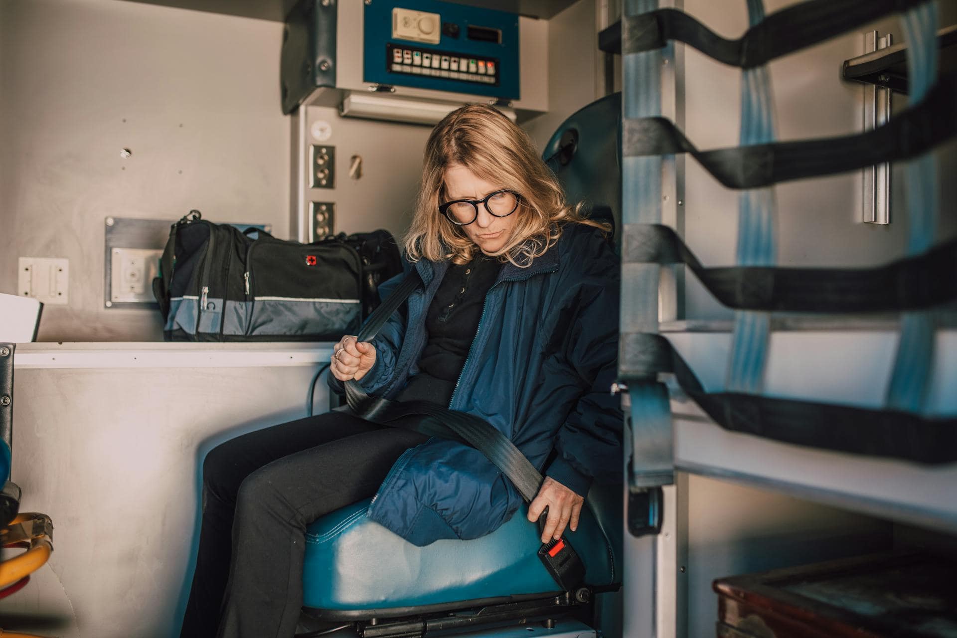 Female paramedic wearing glasses buckling her seatbelt inside an ambulance, preparing for duty.