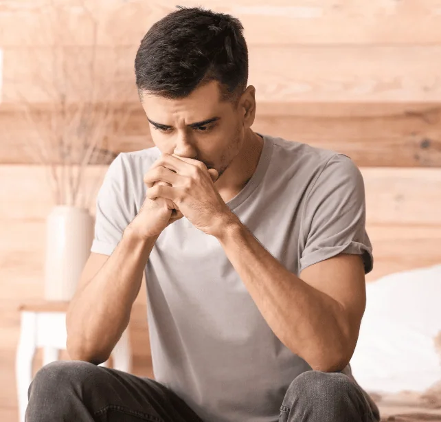 A man sitting on the edge of his bed in a grey t-shirt, hands clasped against his mouth, staring at the floor.
