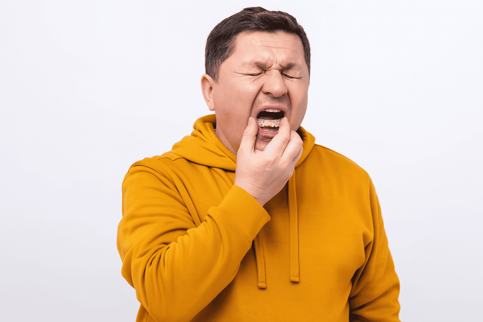 A middle-aged man in a mustard hoodie placing a mouthguard in his mouth with a pained expression, standing against a plain white background.