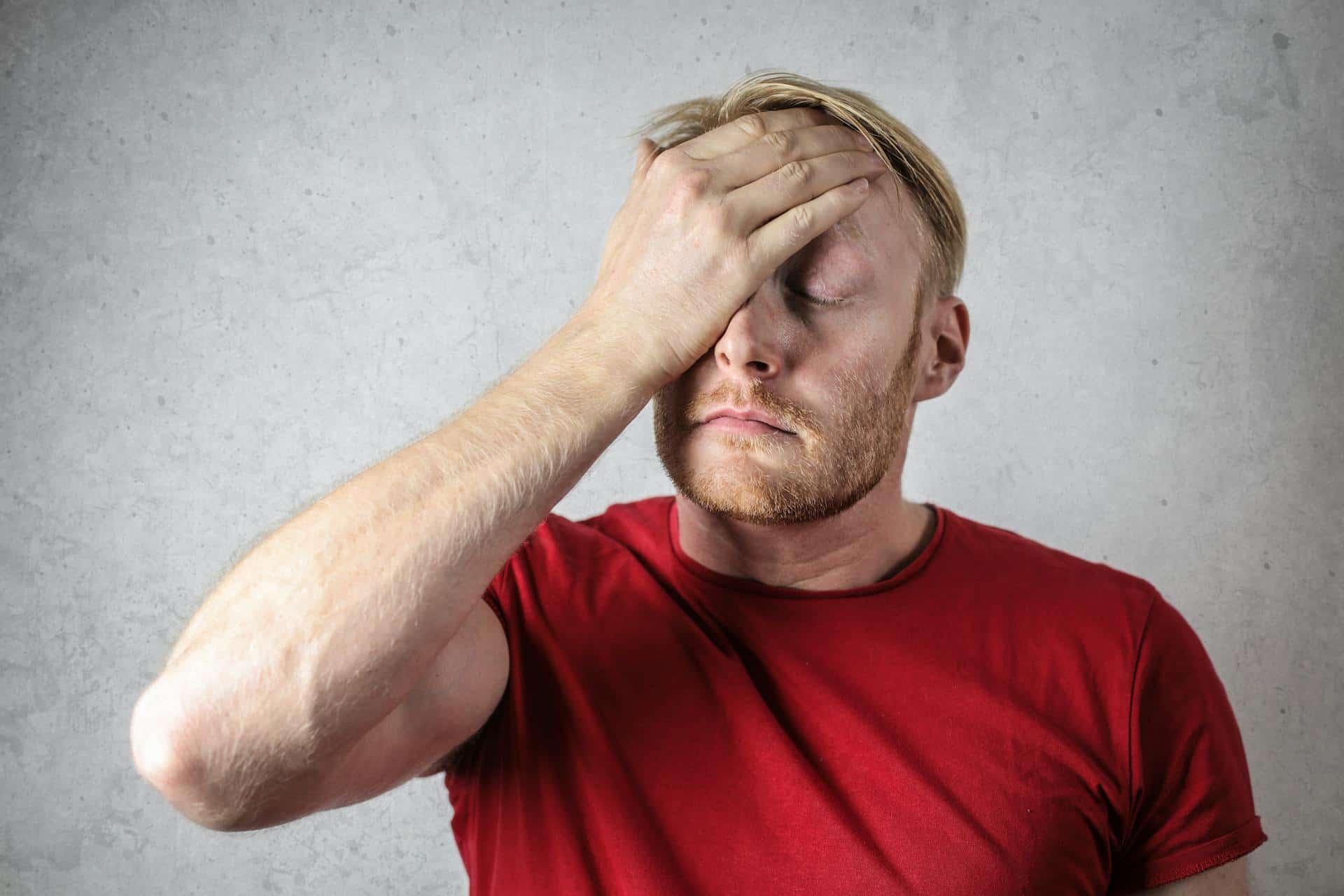 Man in a red shirt with eyes closed, holding his forehead in apparent stress or pain indicating one of the causes of bruxism.