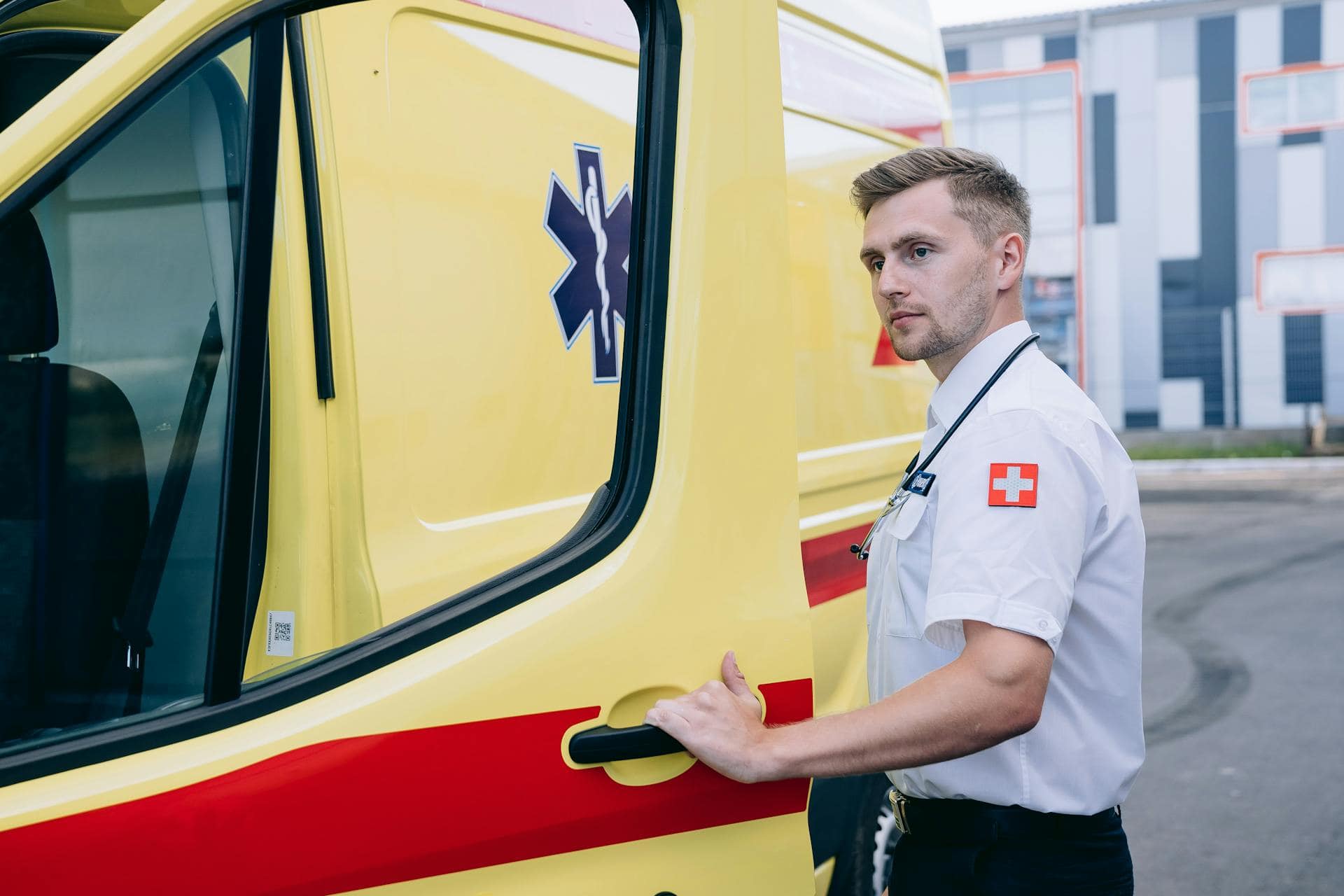 Male paramedic in uniform with a stethoscope standing beside a yellow ambulance, preparing to open the door.