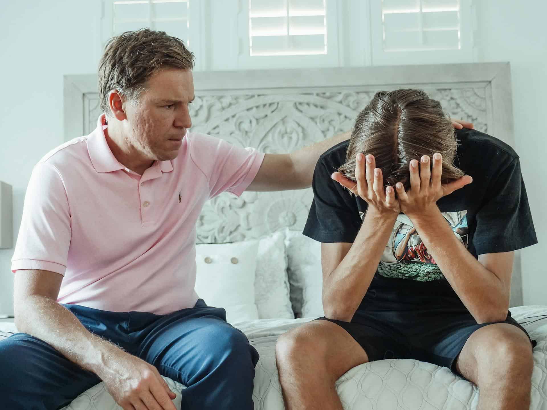 Father comforting his teenage son who appears overwhelmed and stressed, sitting on a bed in a bright room.