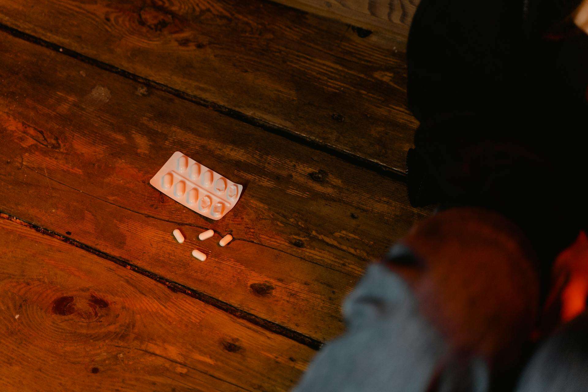 A blister pack and scattered white capsules lie on a wooden floor near a person's legs, suggesting a moment of distress or crisis.