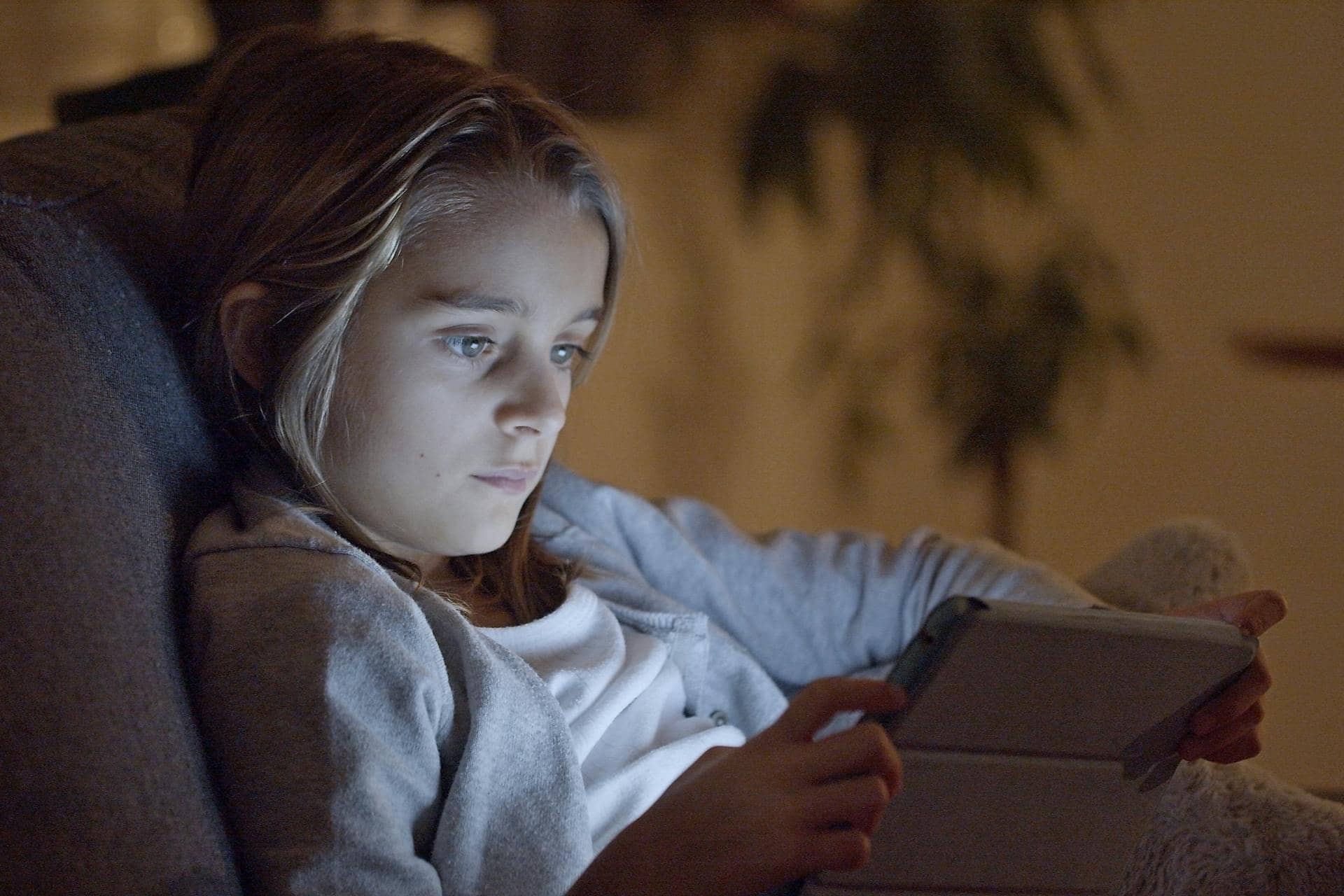 Child with focused expression using a tablet in a dimly lit room, illustrating the link between screens and ADHD.