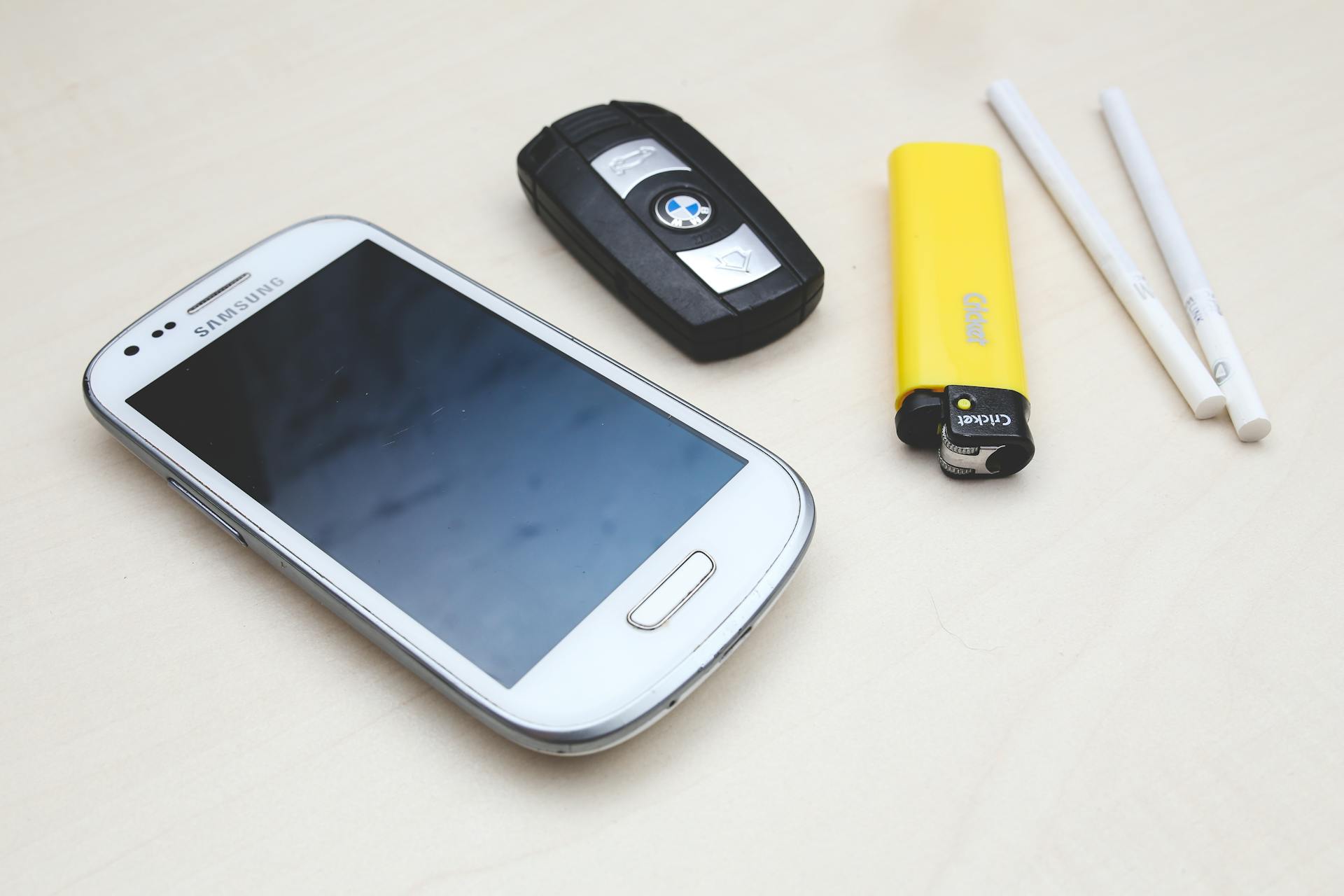 Flat lay of a Samsung smartphone, BMW car key, yellow lighter, and two white cigarette-style objects on a light wooden surface.