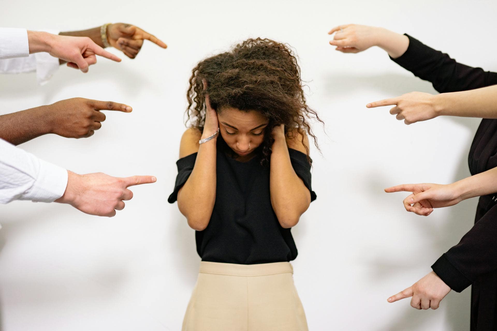 A distressed woman covering her ears while multiple hands point at her, symbolizing social anxiety and fear of judgment.
