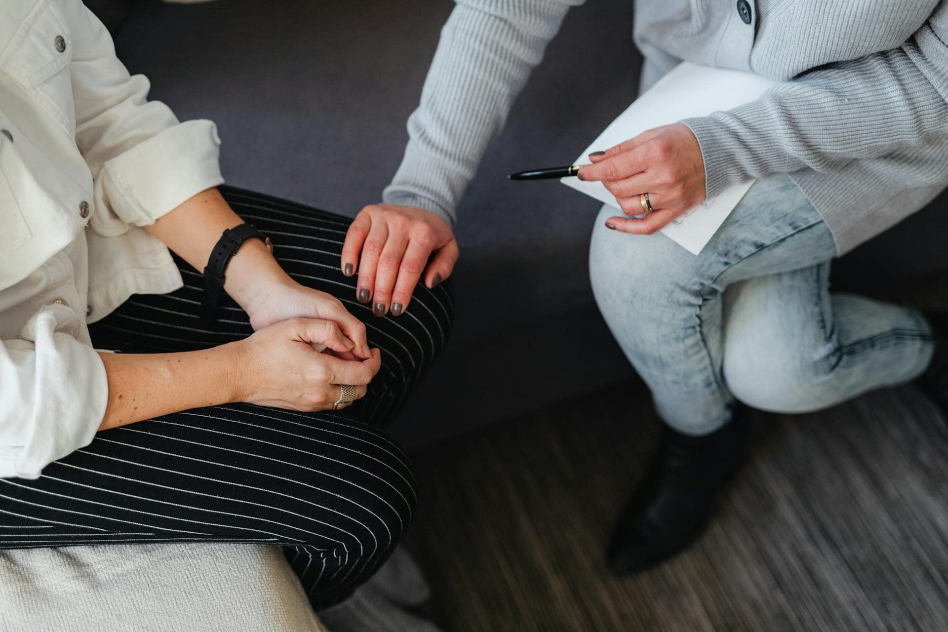A close-up of a supportive therapy session showing two women sitting; one gently places her hand on the other’s knee while holding a pen and notepad, symbolizing trust and care in a therapeutic setting.