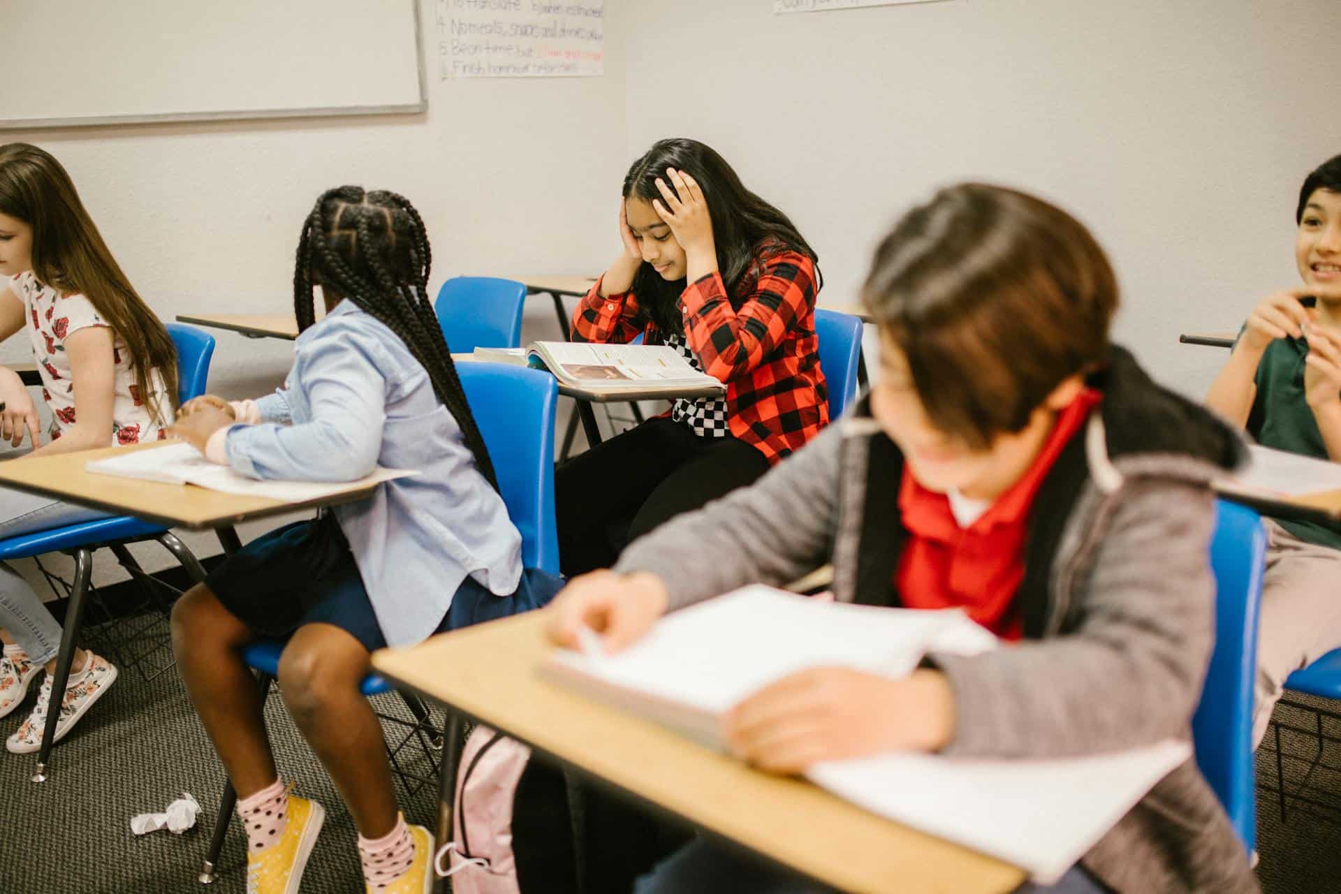 Young student holding her head in frustration while studying in a classroom, surrounded by other classmates focused on their work.
