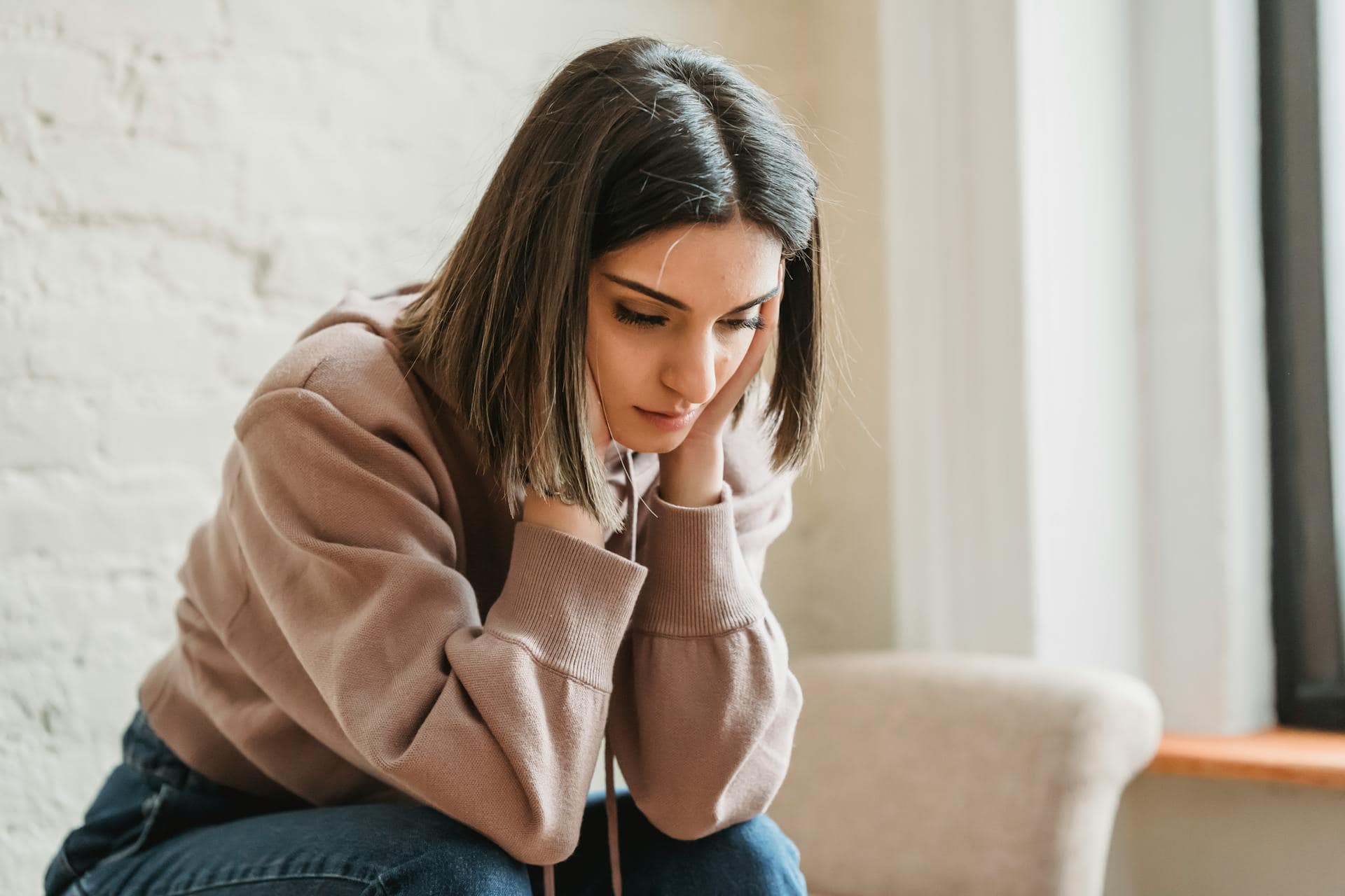 Young woman sitting indoors with a worried expression, holding her face in both hands depicting emotional stress or anxiety.