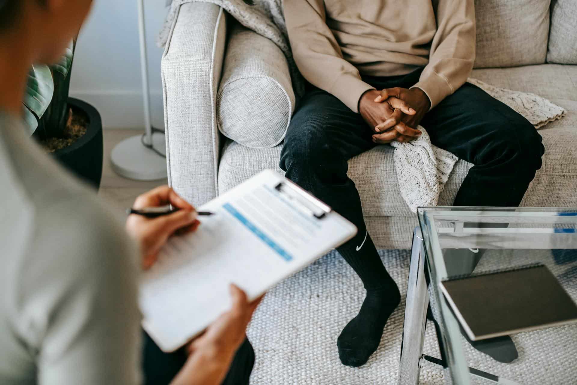 Teen sitting on a couch during a therapy session, with a counselor taking notes on a clipboard across from them.