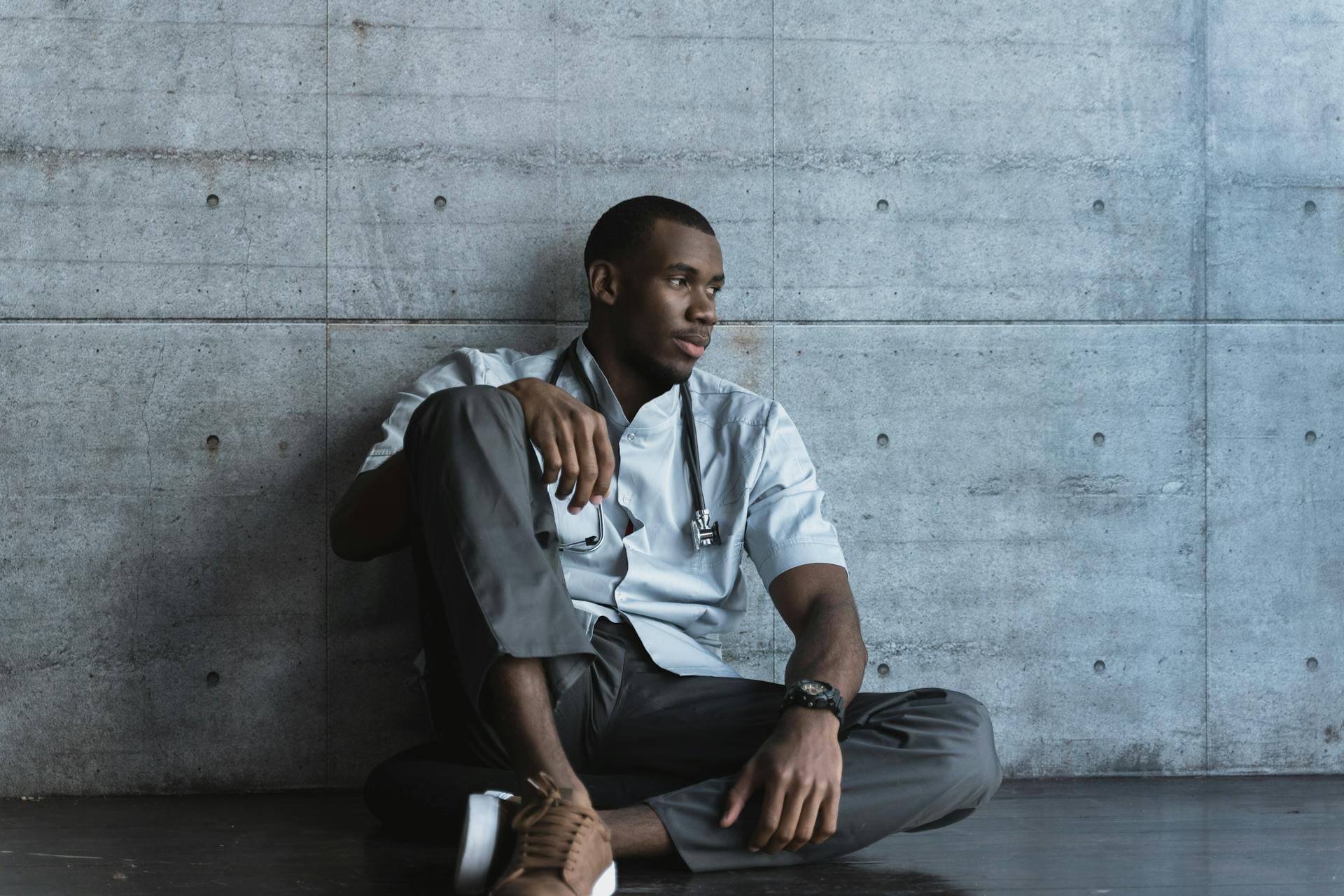 A tired healthcare worker in scrubs and a mask sits on the floor against a wall with eyes closed and head tilted back, portraying physical and emotional exhaustion from long hours and demanding shifts.
