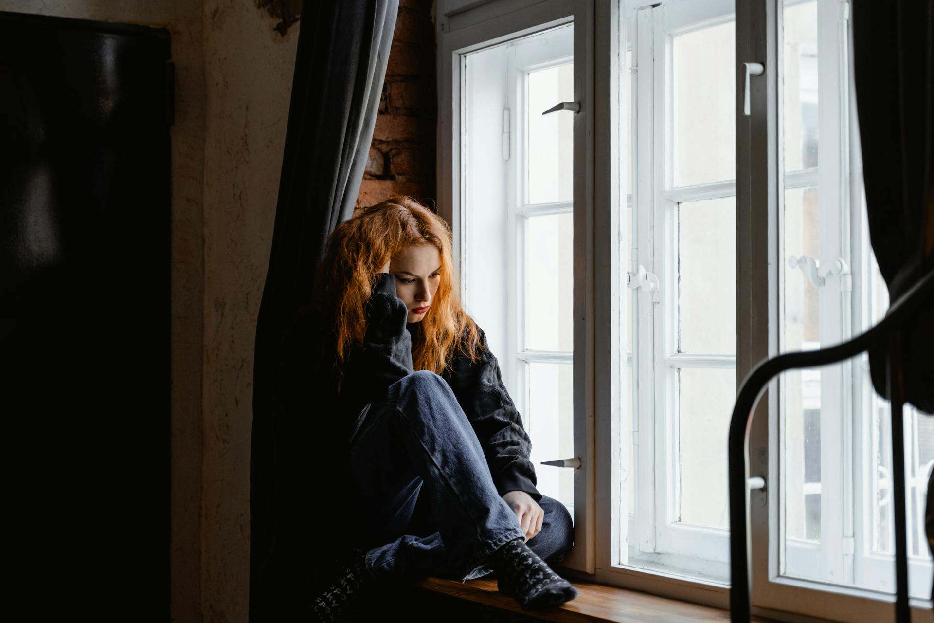 A woman sitting by the window appearing emotionally distressed, possibly experiencing TMJ disorder in women.