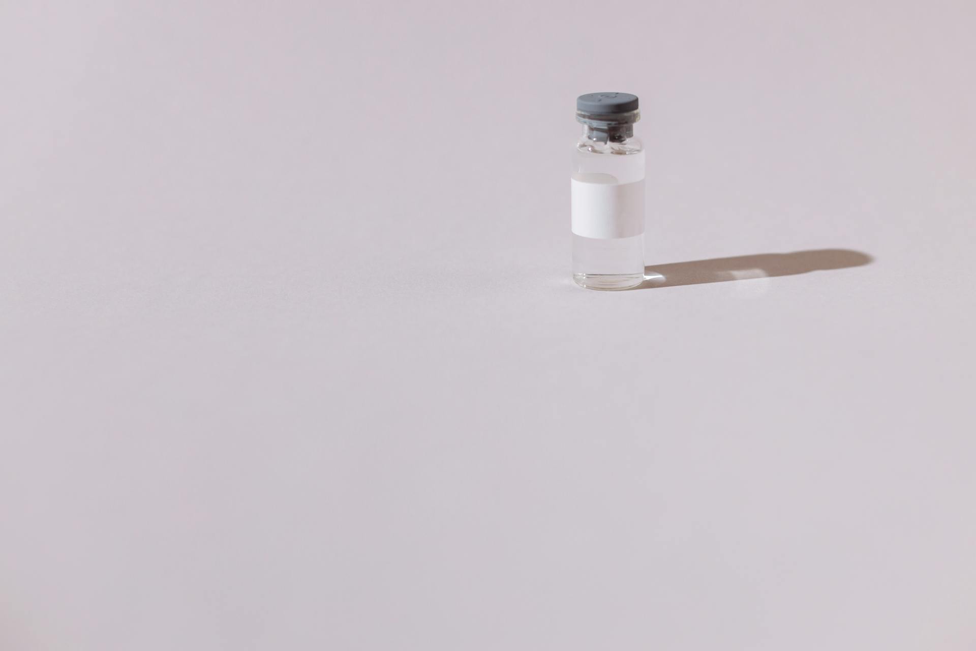 A small glass vial with a blank white label and gray cap, standing on a pale surface with a soft shadow.