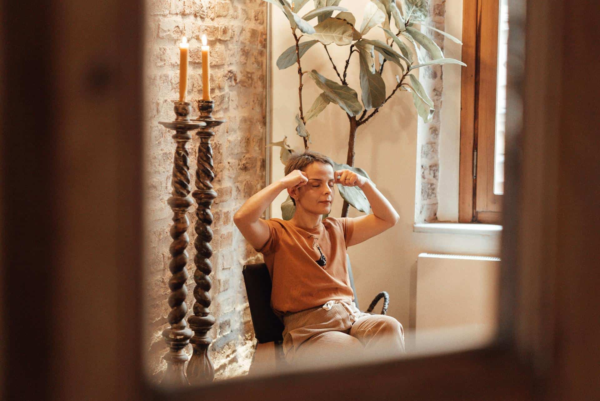 Woman sitting indoors with eyes closed, gently pressing her fingers to her forehead as part of a calming or meditative practice.
