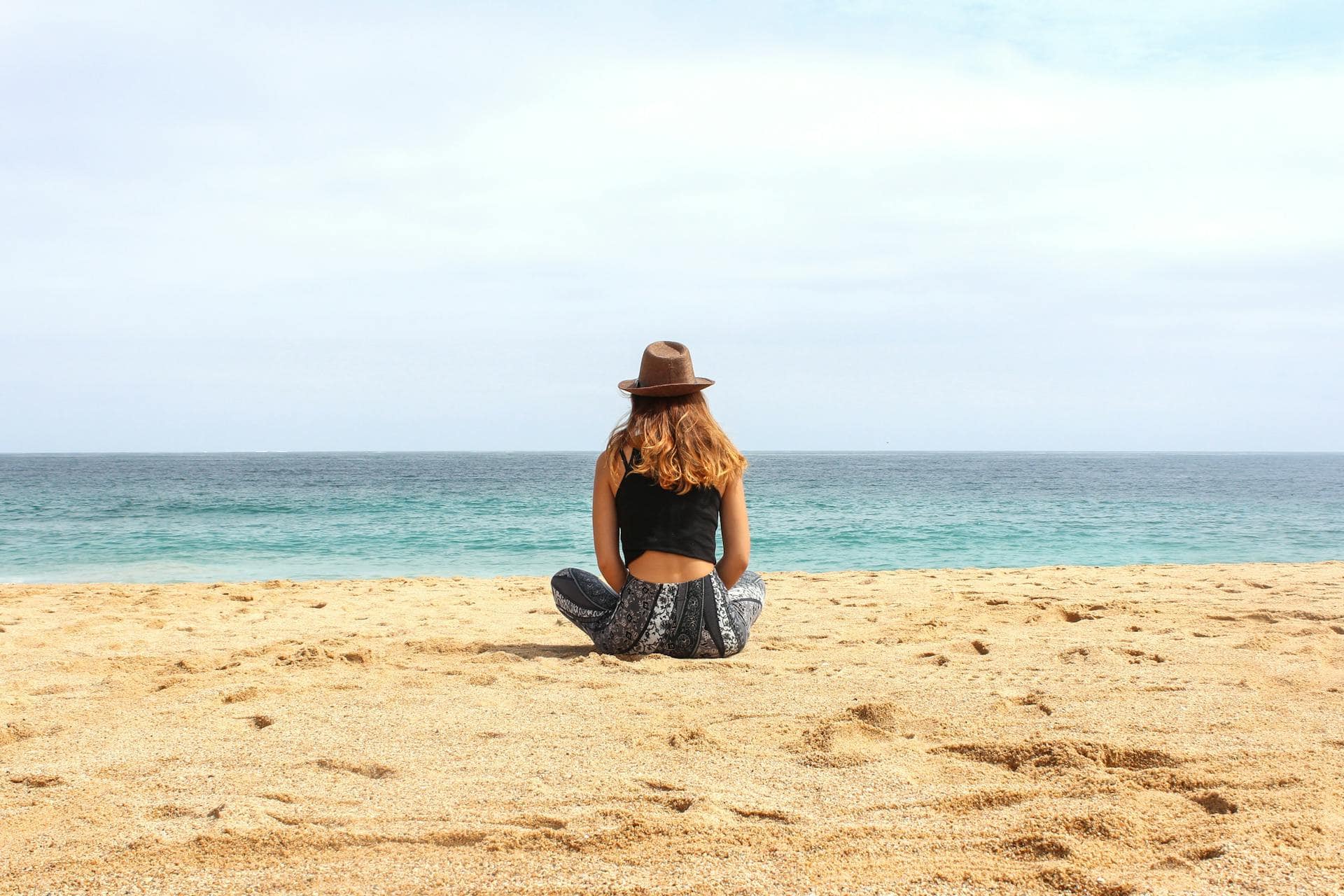 Woman sitting alone on a sandy beach facing the ocean, wearing a hat and relaxed clothing, enjoying a peaceful moment.