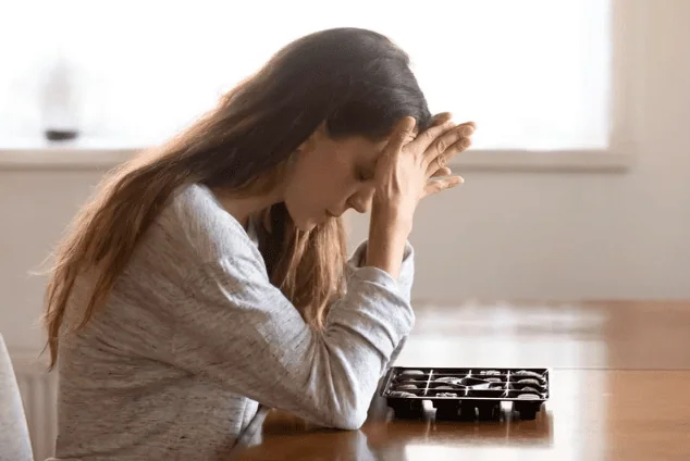 A woman sitting alone at a table with her head down and hands clasped, appearing sad and contemplative