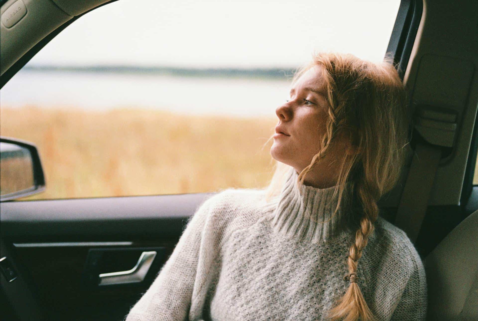 Young woman with a braid in a cozy sweater gazing out of a car window, appearing thoughtful and emotionally distant.