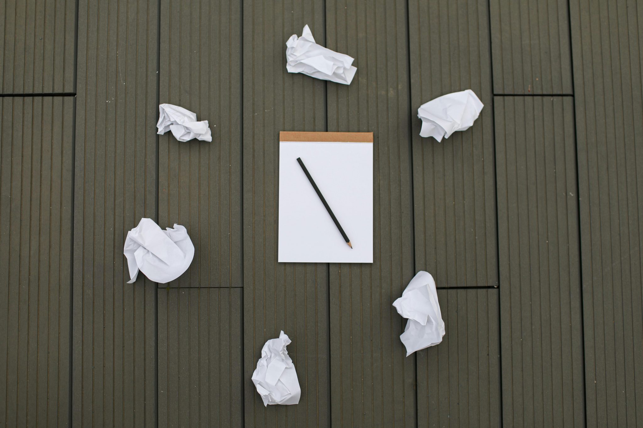 Blank notepad with a pencil surrounded by crumpled paper balls on a wooden surface, symbolising writer’s block or creative frustration.